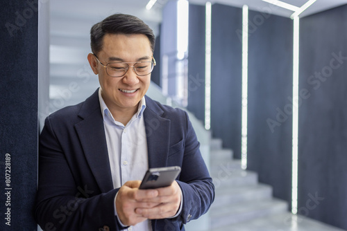 A cheerful Asian businessman in a suit stands using his mobile phone in a contemporary office setting with stylish lighting.