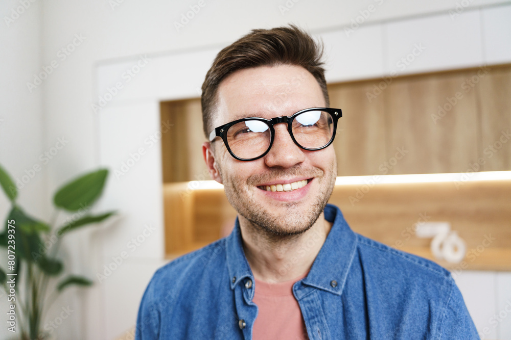 A close-up of a cheerful young man with glasses, smiling brightly in a modern, well-lit office environment. A close-up of a cheerful young man with glasses, smiling brightly in a modern, well-lit office environment.