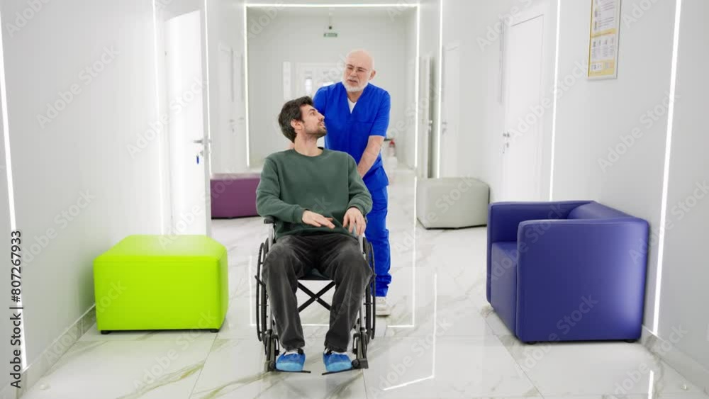 An experienced male doctor in glasses with a gray beard and in a blue uniform communicates with a young brunette guy with stubble in a green jacket who is riding in a chair for the disabled after his An experienced male doctor in glasses with a gray beard and in a blue uniform communicates with a young brunette guy with stubble in a green jacket who is riding in a chair for the disabled after his