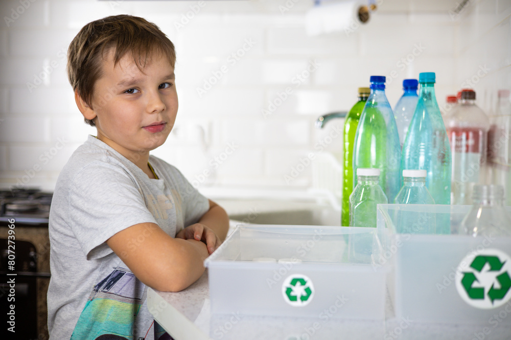 The boy looks thoughtfully at the camera next to boxes with a recycling sign. A child boy in the kitchen sorts plastic for recycling. The child cares about nature. The boy looks thoughtfully at the camera next to boxes with a recycling sign. A child boy in the kitchen sorts plastic for recycling. The child cares about nature.