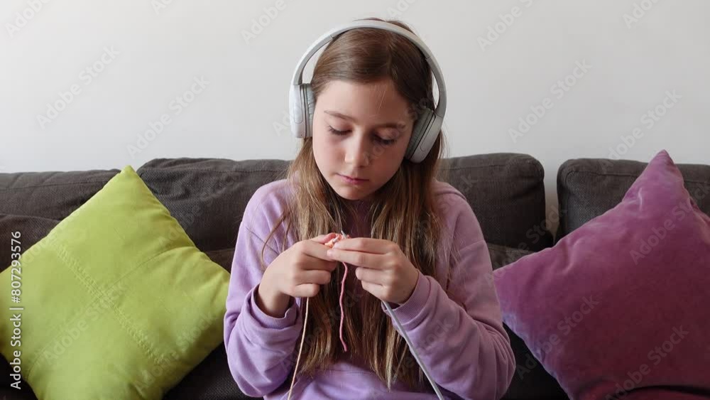 Ten year old girl with headphones knitting colorful wool with needles sitting on a sofa at home