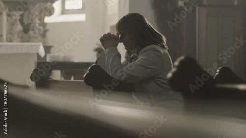 Christian female parishioner sitting on pew bench, praying to God with clenched fists side view medium shot. Mid adult caucasian woman during prayer in empty church photo