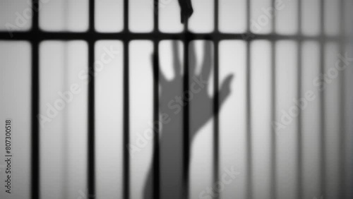 A silhouette of a man's hand appears, its open palm gesturing for help, pleading to stop; trapped within the confines of a bleak prison cell, its image projected onto a white wall.
