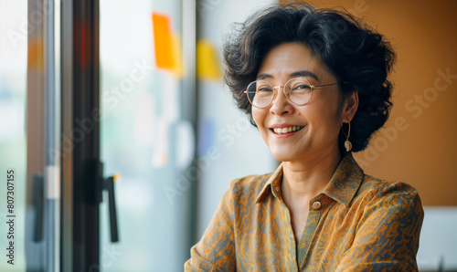 Mature Asian business woman wearing glasses looking out at her modern office. Planning strategy and team. Executive owner, CEO, retirement planning, financial plan. 