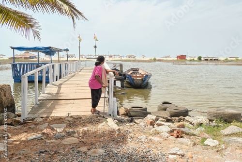 A senior African American woman with walking stick standing on a pier to a sandy beach with blue sky and canoe in the background, Ghana