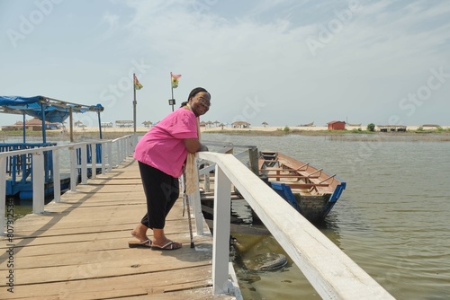 A senior African American woman with walking stick standing on a pier looking and smiling towards camera with a sandy beach with blue sky and canoe in the background, Ghana