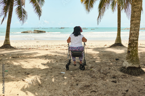 Senior African American woman sitting on a mobile rollator walker under palm trees at golden sandy Abandze beach looking at the blue ocean and fishing boats, Ghana