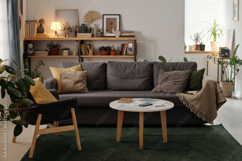 Fototapeta premium Low round table with mobile gadgets, financial bills and notepad standing in the center of living room against couch with cushions