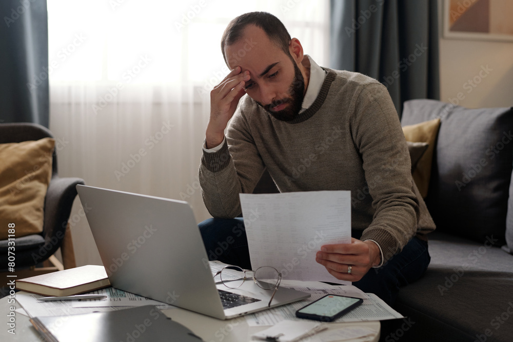 Young tense man with beard holding financial bill in front of himself and checking data while sitting on couch in front of laptop Young tense man with beard holding financial bill in front of himself and checking data while sitting on couch in front of laptop