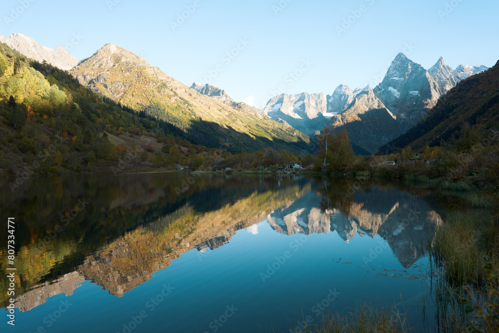 A picturesque mountain lake with aquatic plants in the water near the shore. Rocky snow-capped mountains and autumn trees are reflected in the water surface.