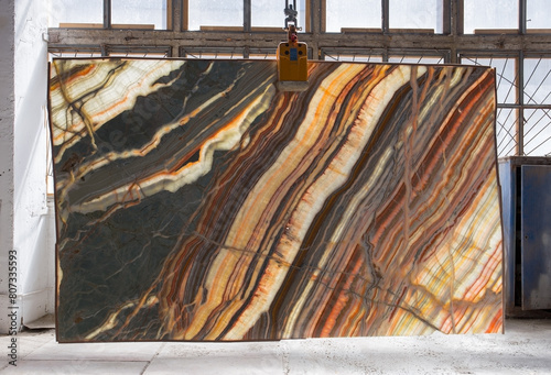 Picture of a backlit onyx slab on a hanger, shot in natural light, a cut of decorative marble in a warehouse, a large piece of marble. High resolution photo. Onyx Rainbow