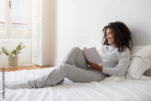 A Hispanic woman in casual clothing sitting on a neatly made bed, engrossed in writing in diary. The room appears cozy with soft lighting, and there are a few pillows scattered on the bed.