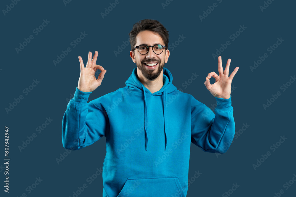 A cheerful man with a beard and glasses is wearing a bright blue hoodie and making the OK sign with both hands, suggesting positivity and approval. He is standing against a monochrome blue background A cheerful man with a beard and glasses is wearing a bright blue hoodie and making the OK sign with both hands, suggesting positivity and approval. He is standing against a monochrome blue background