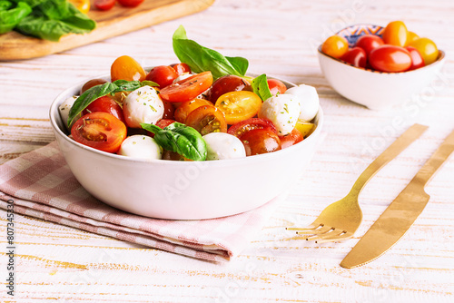 Fresh juicy homemade caprese salad with baby mozzarella cheese, cherry tomatoes, basil leaves and olive oil on white wooden table with colden cutlery set and napkin. Traditional Italian cuisine. photo