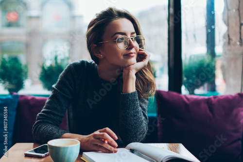 Dreaming beautiful hipster girl looking away while resting at cafeteria and writing romantic essay, attractive young woman in eyeglasses enjoying free time and planning to do list in notebook photo
