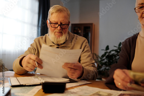 Serious mature man with white beard holding financial document with housing payment sums and looking through information
