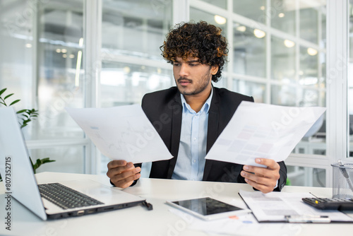 Fotografi Focused young businessman with curly hair wearing a suit examining multiple documents and reports while sitting at a modern office desk