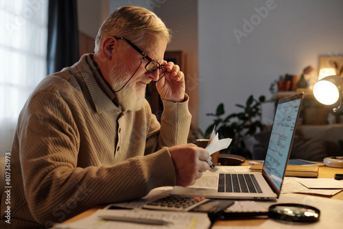 Side view of serious elderly man in eyeglasses checking payment information on receipts while sitting in front of laptop photo