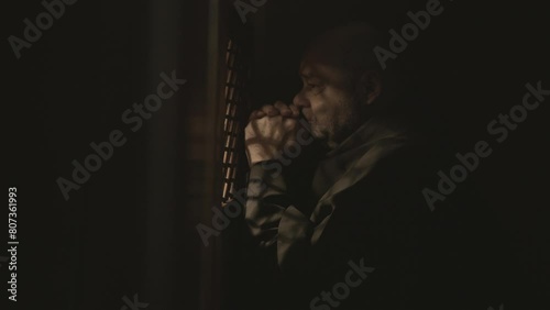 Mid adult monk sitting in confessional booth and praying for forgiveness. Caucasian bald Christian clergyman listening to confession, crossing and speaking