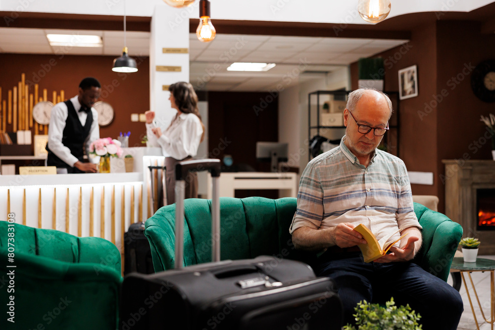 Obraz premium Speaking to young concierge at main desk of luxury resort is caucasian female traveller. While old woman completes booking process at counter an elderly man reading a book relaxes in hotel lobby.