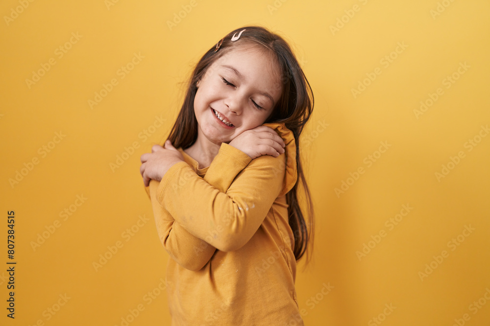 Adorable hispanic girl with a joyful smile standing and hugging herself on isolated yellow background, exuding confidence and positive self love Adorable hispanic girl with a joyful smile standing and hugging herself on isolated yellow background, exuding confidence and positive self love