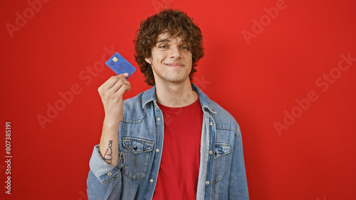 Smiling young man with curly hair holding credit card against red background photo