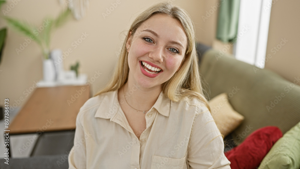 Smiling blonde woman in a casual beige shirt posing comfortably at her modern furnished living room. Smiling blonde woman in a casual beige shirt posing comfortably at her modern furnished living room.