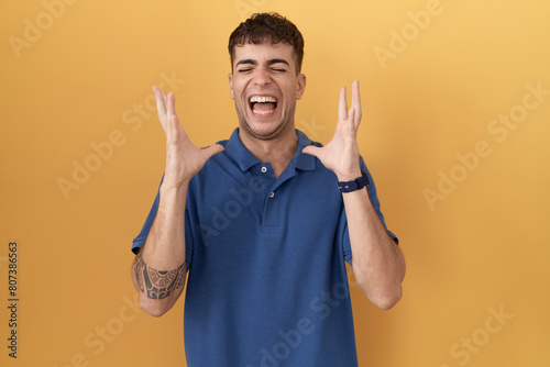 Young hispanic man standing over yellow background celebrating mad and crazy for success with arms raised and closed eyes screaming excited. winner concept