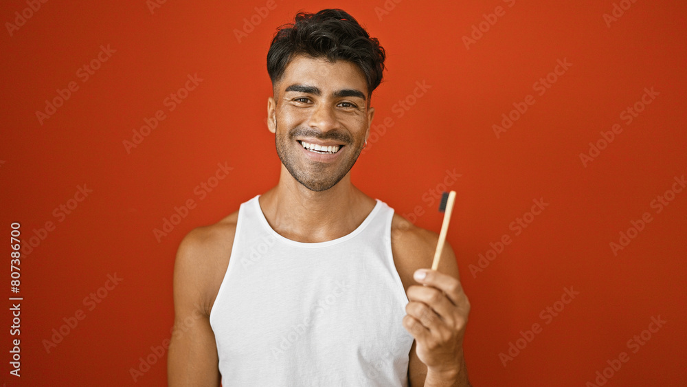 A cheerful young hispanic man with a beard poses in a sleeveless shirt against an isolated red background, holding a pencil. A cheerful young hispanic man with a beard poses in a sleeveless shirt against an isolated red background, holding a pencil.