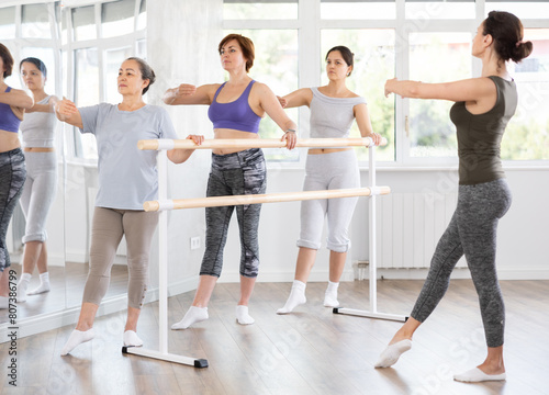 Elderly woman carefully executing battement tendu at barre in beginners ballet class guided by female instructor in sunlit choreography studio. Concept of active healthy hobby for seniors.. photo