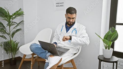 Bearded man in lab coat checks watch while working on laptop in modern clinic lobby with free wifi sign. photo