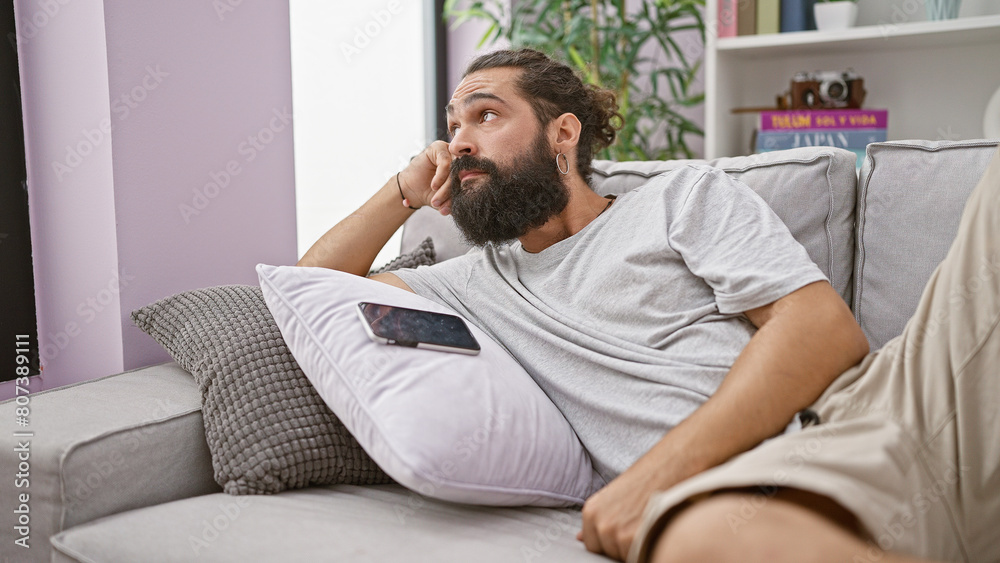 Thoughtful bearded man lounging on couch with smartphone, expressing relaxation and comfort in a cozy room. Thoughtful bearded man lounging on couch with smartphone, expressing relaxation and comfort in a cozy room.
