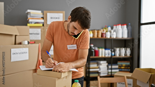 Young hispanic man multitasking, talking on phone while volunteering at a donation center, surrounded by cardboard boxes