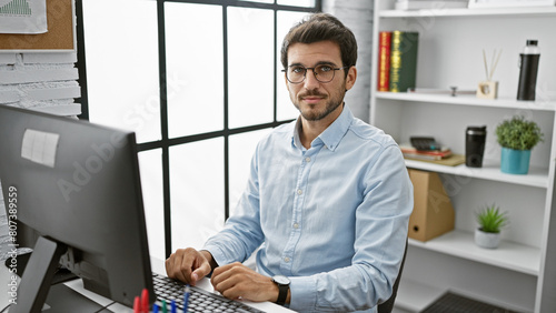 A young hispanic man with glasses and a beard poses in a modern office setting, exuding confidence and professionalism.