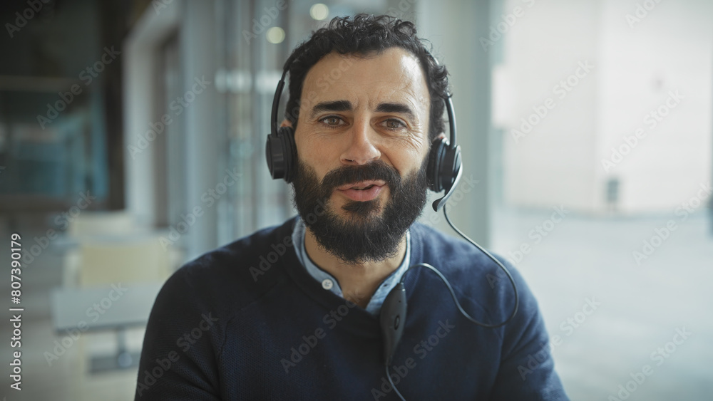 Naklejka premium Handsome bearded man wearing headphones smiling in a bright modern office setting.