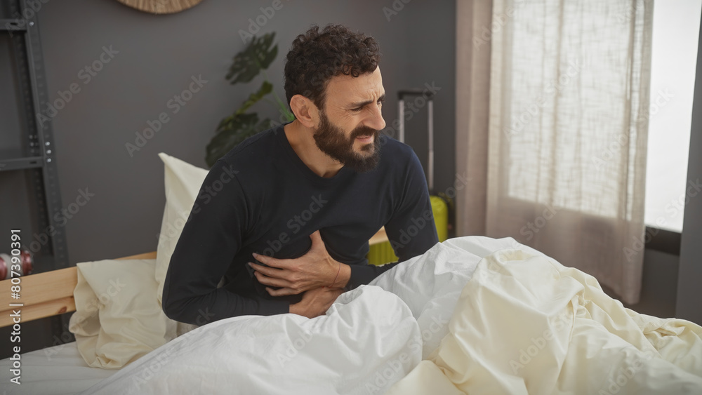 A bearded man in pain sitting on a bed indoors, clutching his stomach, evoking feelings of sickness or discomfort in a bedroom setting. A bearded man in pain sitting on a bed indoors, clutching his stomach, evoking feelings of sickness or discomfort in a bedroom setting.