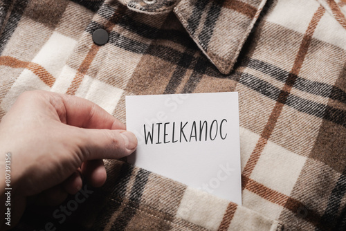  White card with a handwritten inscription "Wielkanoc", held in the hand against the background of a brown plaid shirt (selective focus), translation: easter