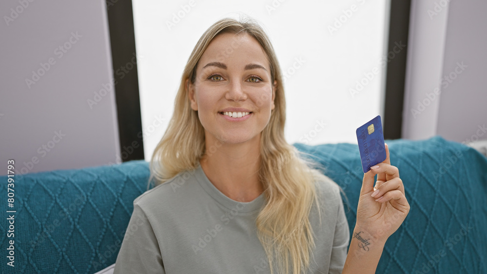 Blonde woman smiling holding credit card in modern home interior. Blonde woman smiling holding credit card in modern home interior.