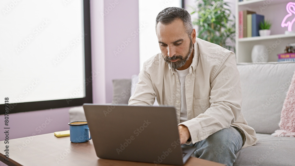 A mature bearded man works on a laptop in a cozy living room with a sofa and home decor. A mature bearded man works on a laptop in a cozy living room with a sofa and home decor.