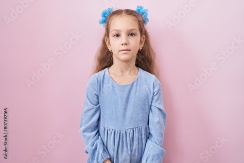 Young little girl standing over pink background with serious expression on face. simple and natural looking at the camera.