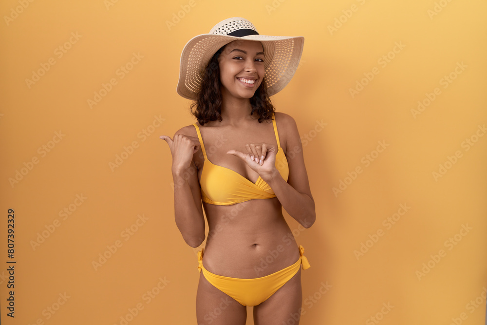 Young hispanic woman wearing bikini and summer hat pointing to the back behind with hand and thumbs up, smiling confident Young hispanic woman wearing bikini and summer hat pointing to the back behind with hand and thumbs up, smiling confident