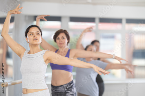 Women of different ages exercise near the ballet barre at group training session in the batman batyu position