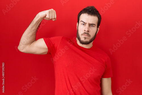 Young hispanic man wearing casual red t shirt strong person showing arm muscle, confident and proud of power