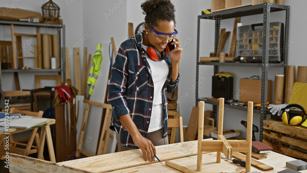 African american woman talking on the phone while measuring wood in a carpentry workshop.