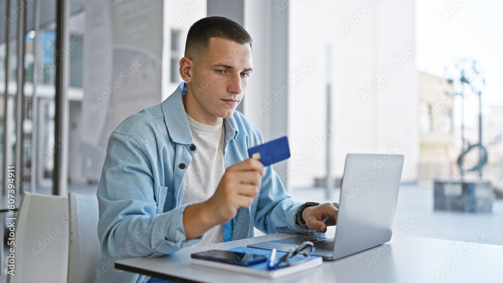 Hispanic man holding credit card while using laptop in modern indoor university setting