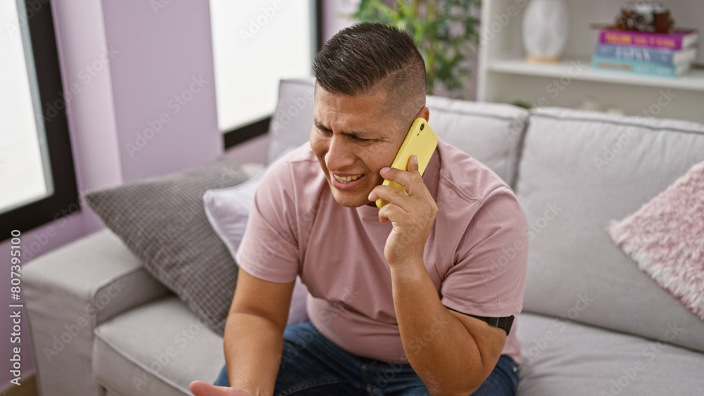 Handsome, worried latin guy in serious phone talk, upset at home. unhappy young man speaking on smartphone, sitting on living room sofa, deep in conversation, addressing problems