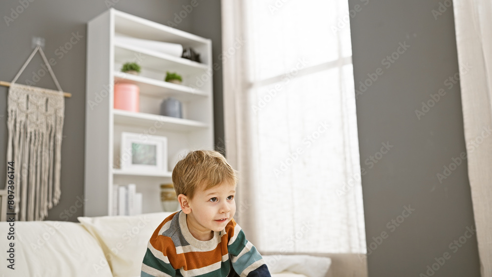Blonde toddler boy sitting thoughtfully on a bed inside a well-lit, cozy bedroom. Blonde toddler boy sitting thoughtfully on a bed inside a well-lit, cozy bedroom.