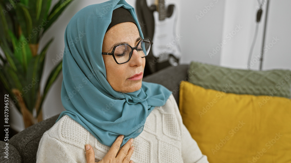 Middle-aged muslim woman in hijab relaxing in living room with eyes closed and serene expression.