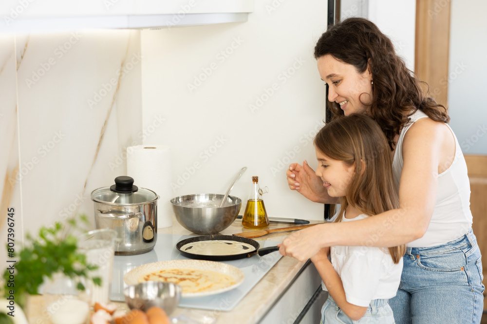 Cheerful middle-aged woman preparing crepes in frying-pan together with her daughter at home