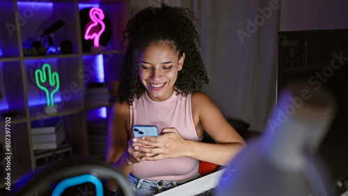 Smiling young hispanic woman streamer, a beautiful gamer thriving in her dark, neon-lit gaming room, confidently streaming via her smartphone